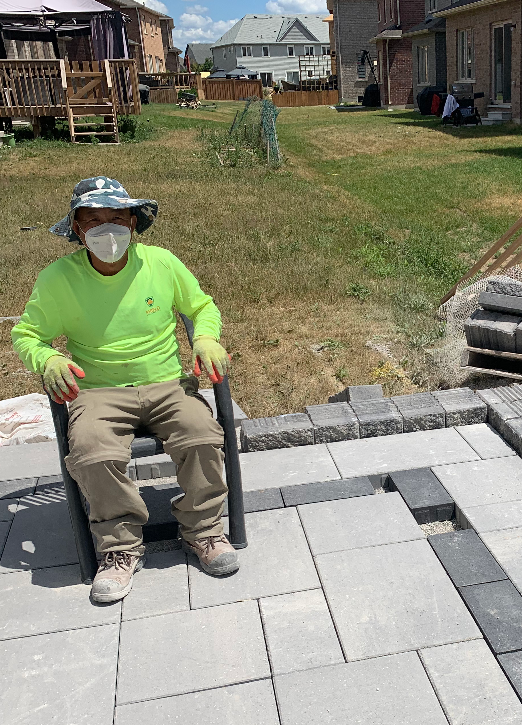 Person wearing a face mask and hat sitting on a chair in a backyard patio under construction, with houses and a grassy area in the background.