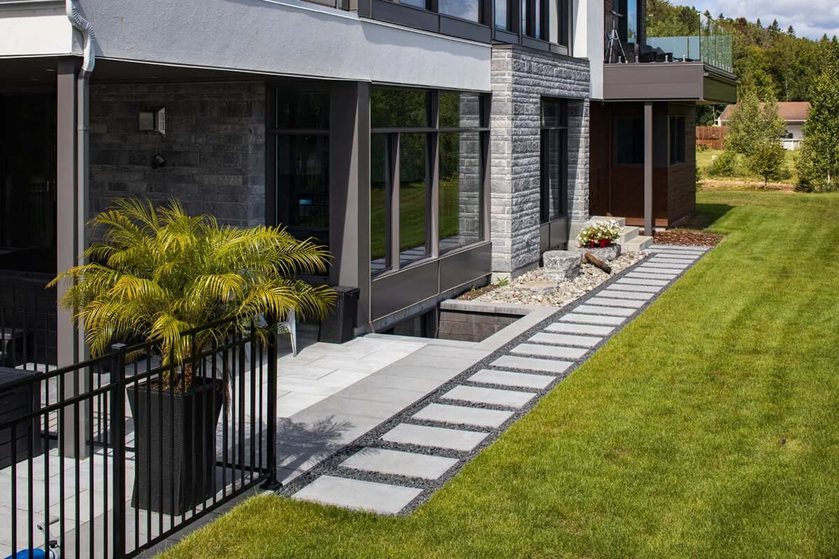 Modern house exterior with stone and glass facade, potted palm, patio, stone path, and green lawn.