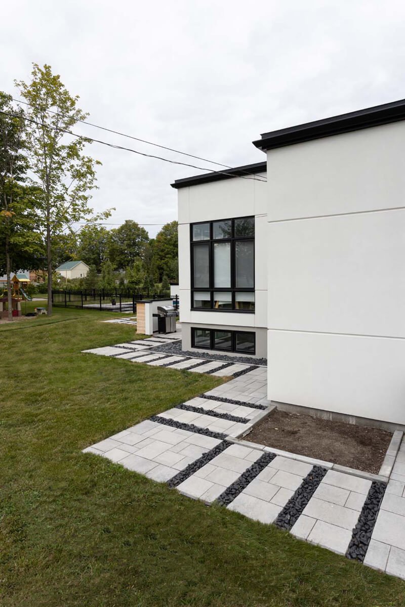 Modern house exterior with large windows, a paved pathway with gravel accents, and surrounding grass and trees.