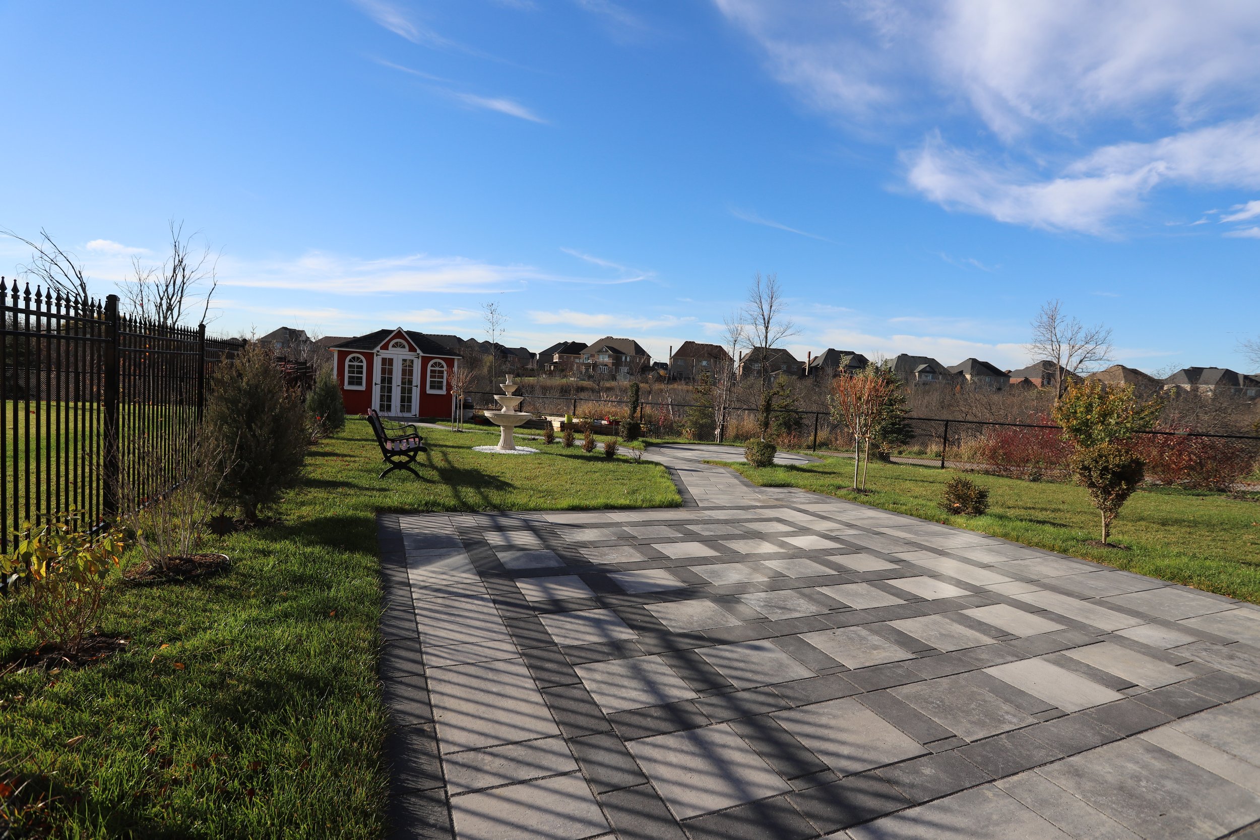 A landscaped backyard with a paved patio, a red garden shed, green grass, a bench, a fountain, and a fence. Sunny sky and residential houses in the background.