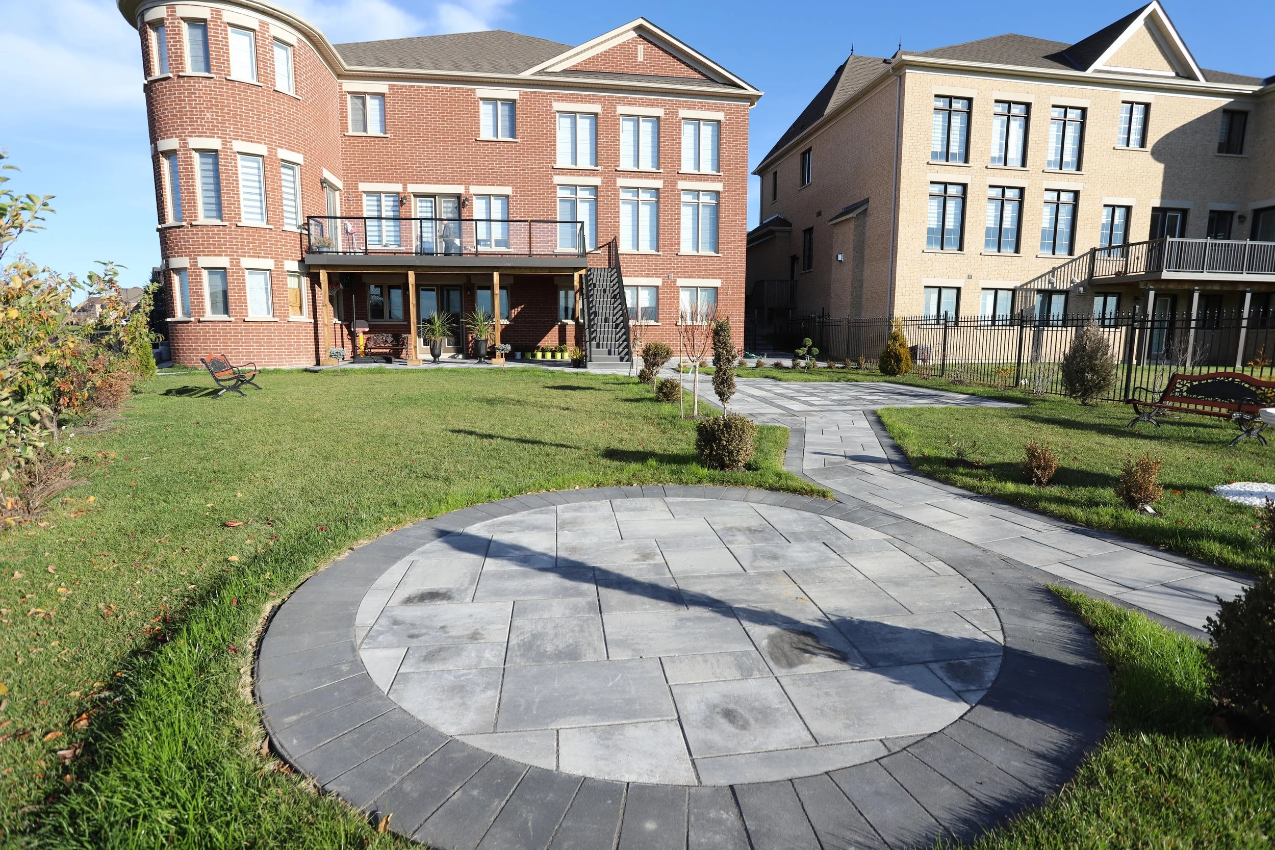 Brick residential building with circular stone walkway and landscaped garden featuring benches, bushes, and small trees.