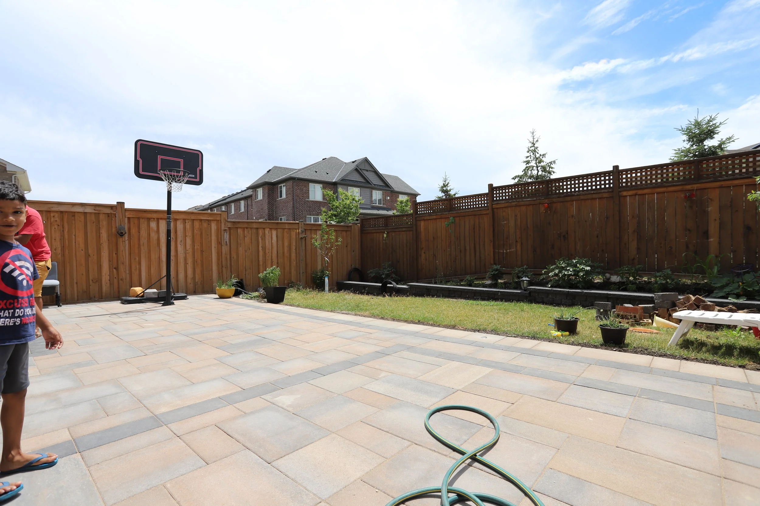 Backyard with basketball hoop on patio, garden area along wooden fence, and a garden hose on the ground. Part of a house and a person visible on the left.