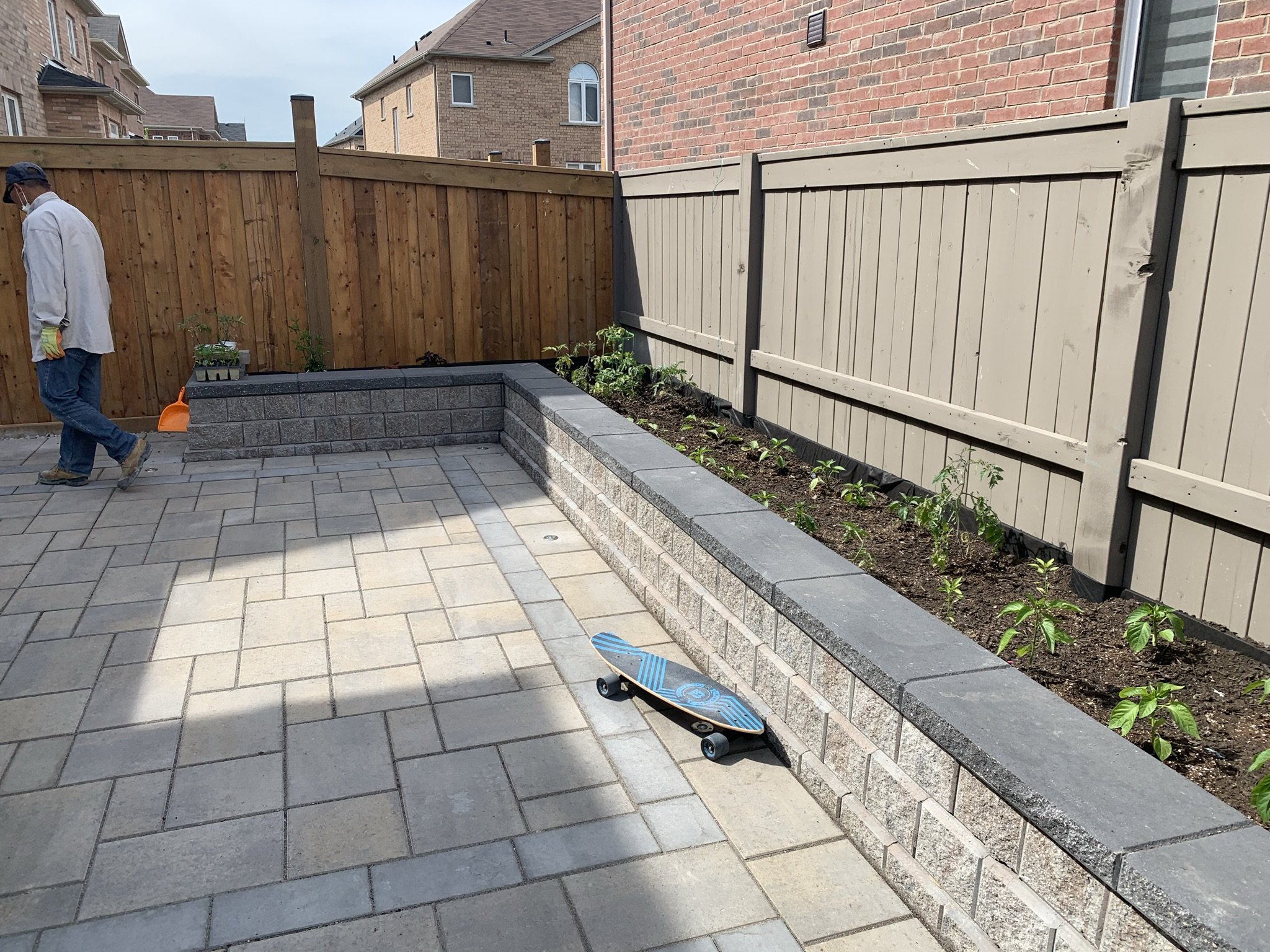 Backyard patio with paver stones, elevated garden bed, skateboard on the ground, and a person walking along a wooden fence.