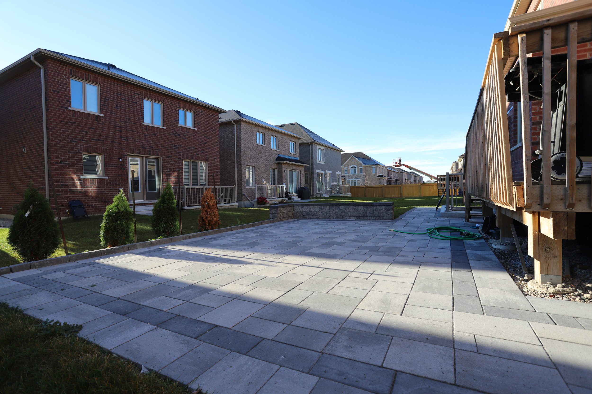 A residential backyard with a paved patio area, surrounded by brick houses, a wooden deck, small trees, and a garden hose on the ground.