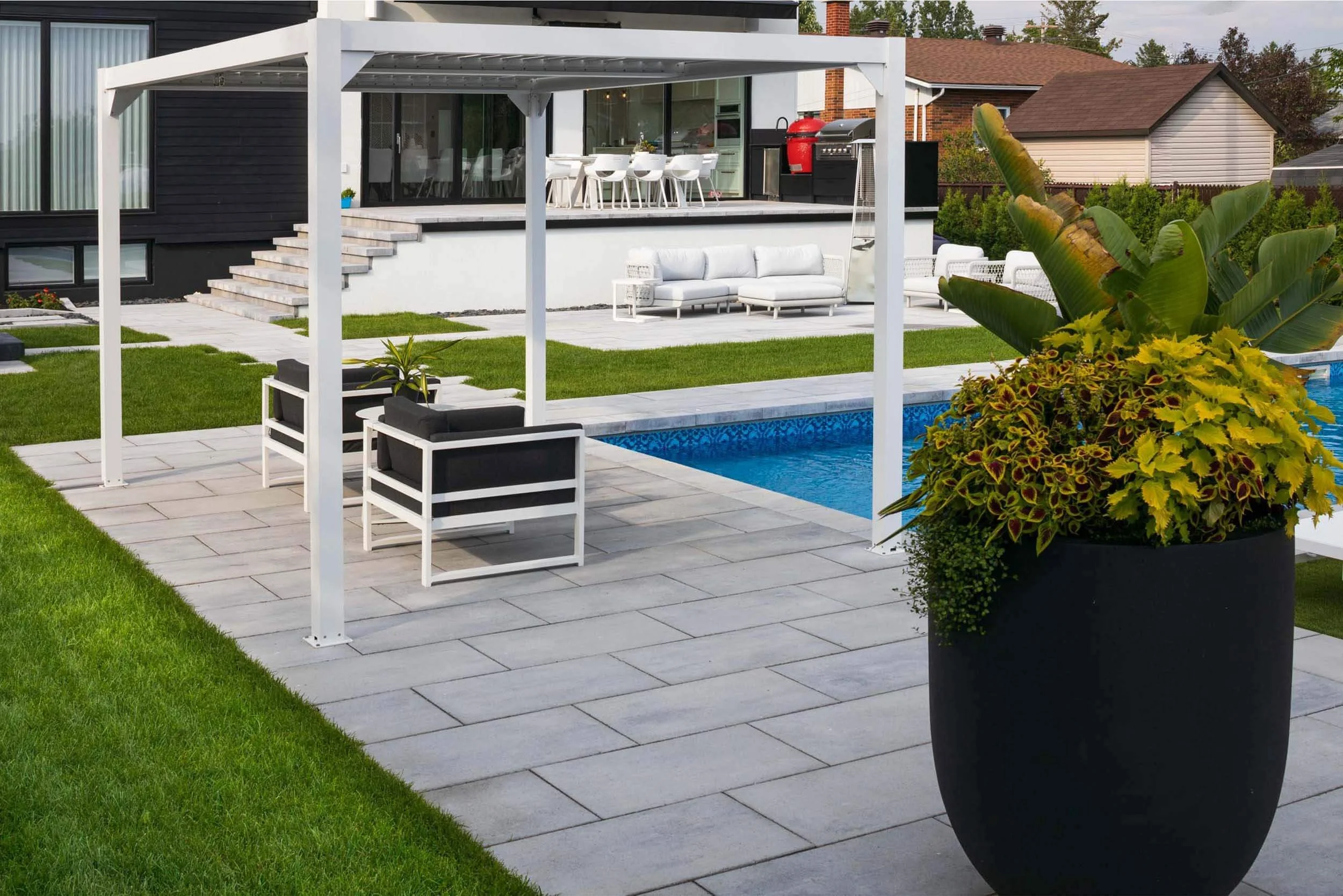 Modern backyard with a swimming pool, outdoor seating under a pergola, manicured lawn, and large potted plants.
