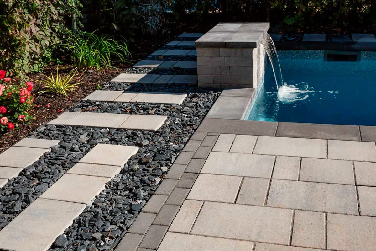 Outdoor pool with stone paving and a water feature cascading into the pool. Pathway with stepping stones and decorative gravel is surrounded by plants and flowers.