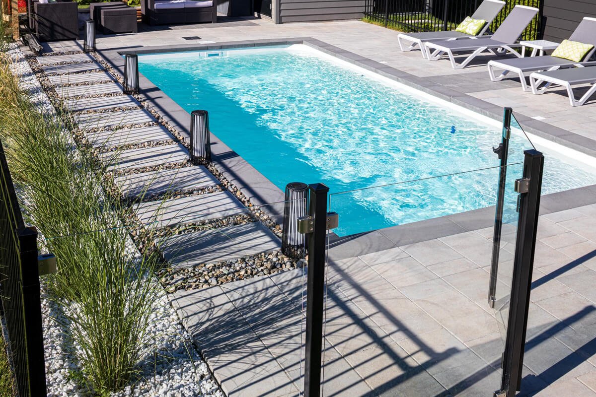 Modern backyard with a rectangular swimming pool surrounded by glass fencing, contemporary lounge chairs, and a landscaped area with decorative grass and stone path.