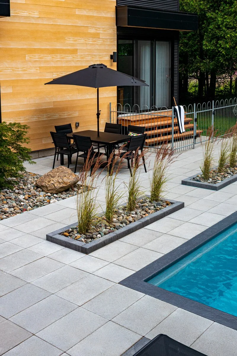Modern backyard patio with rectangular pool, patio furniture, black umbrella, and recessed plant beds. View of a yellow wooden house facade, with stairs and a towel draped over a railing.