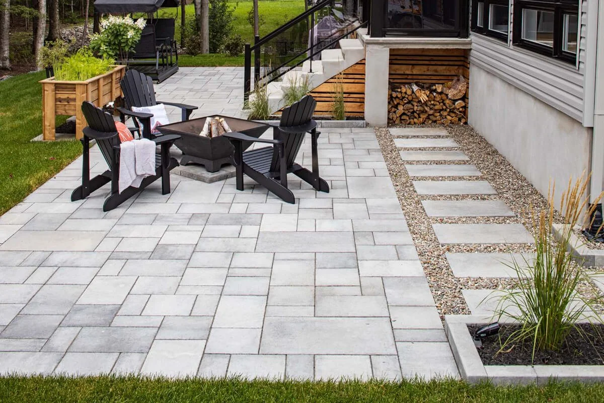 Outdoor patio with gray pavers, Adirondack chairs, a fire pit, wood storage, and landscaped surroundings.