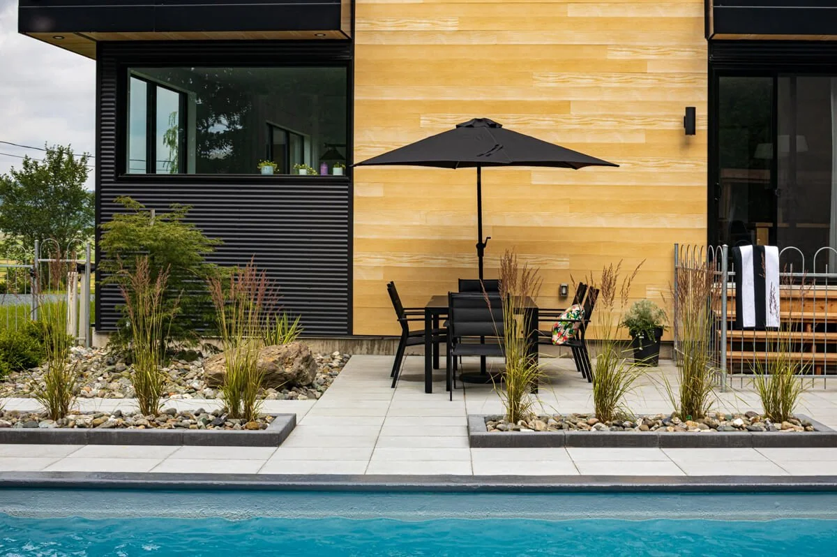 Modern patio with table, chairs, and umbrella next to a swimming pool, alongside landscaped garden with decorative grasses and rocks.