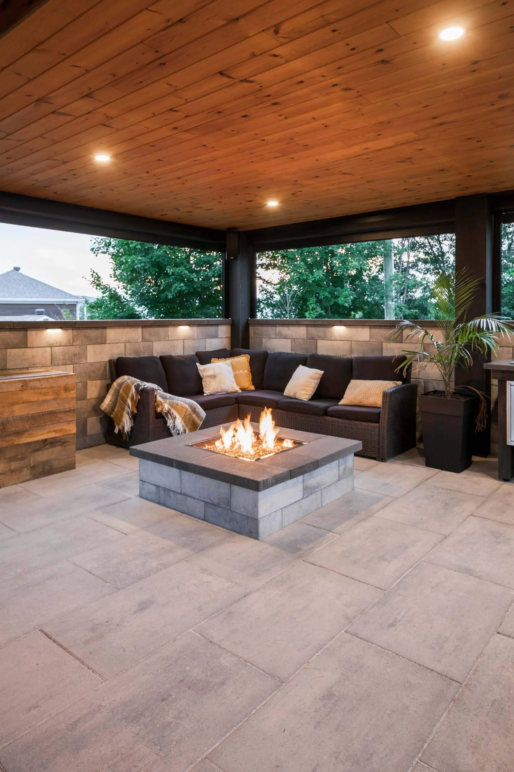 Covered patio with modern fire pit, sectional sofa, wooden ceiling, and plants.