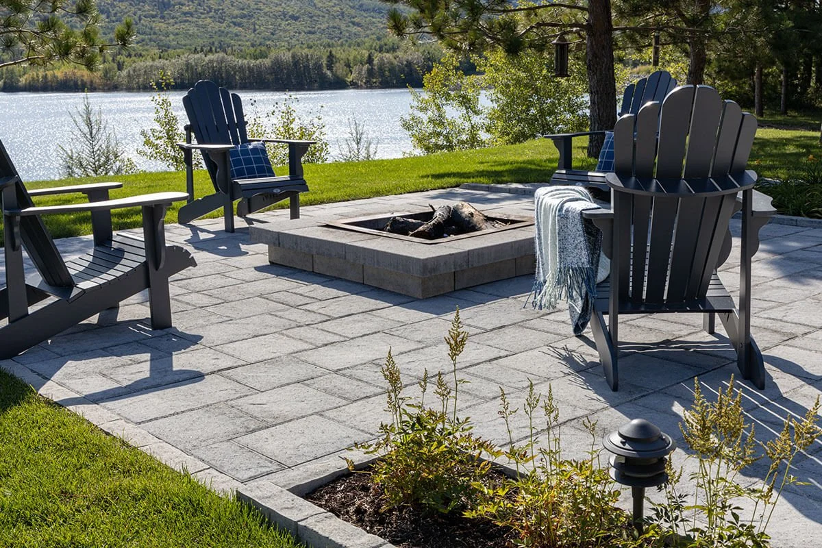 Patio with Adirondack chairs around a fire pit overlooking a lake and trees in the background.