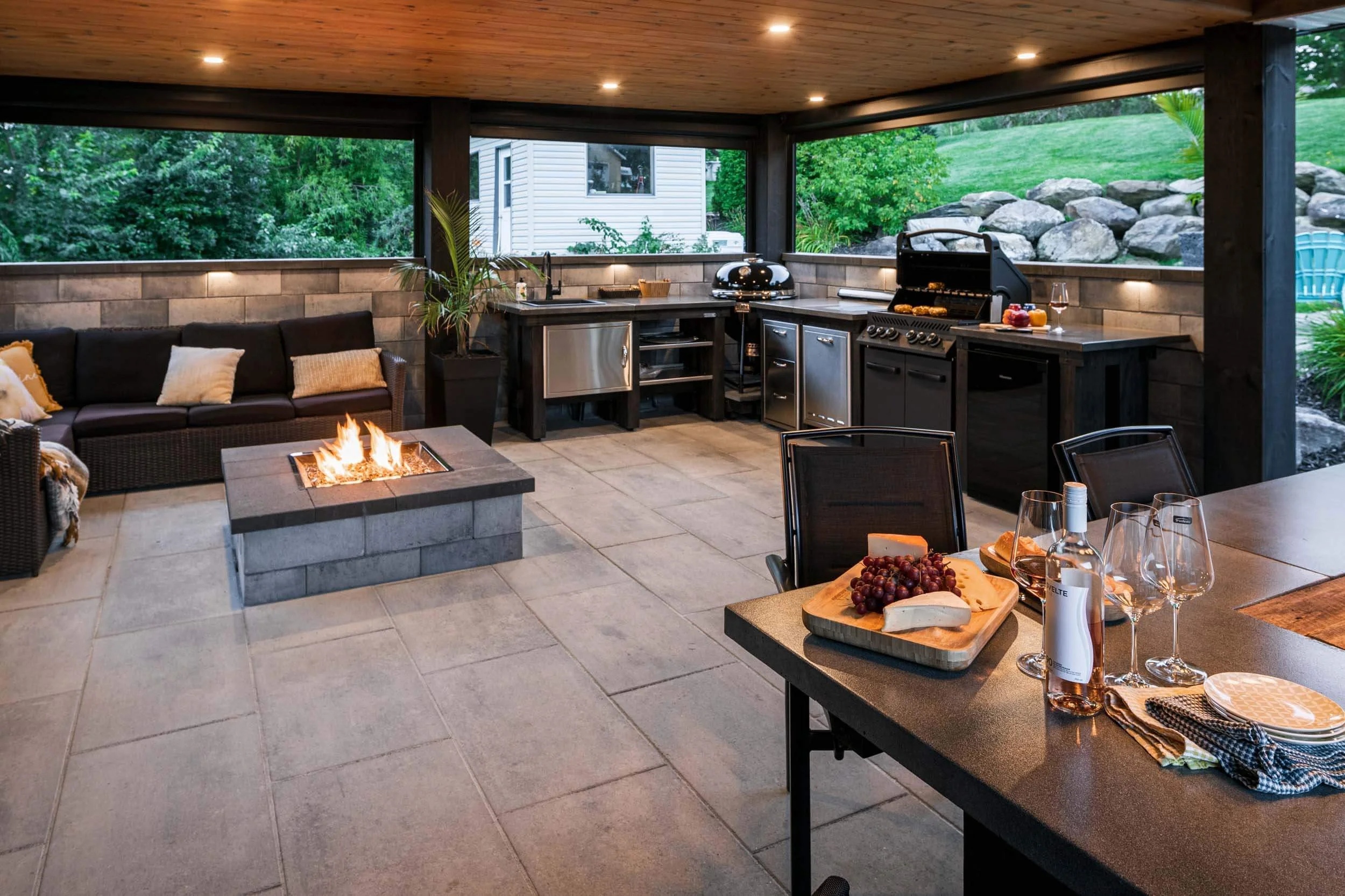 Covered patio with outdoor kitchen, featuring a fire pit, grill, sink, countertop, sofa, dining table with cheese and wine, stone flooring, and surrounding greenery.