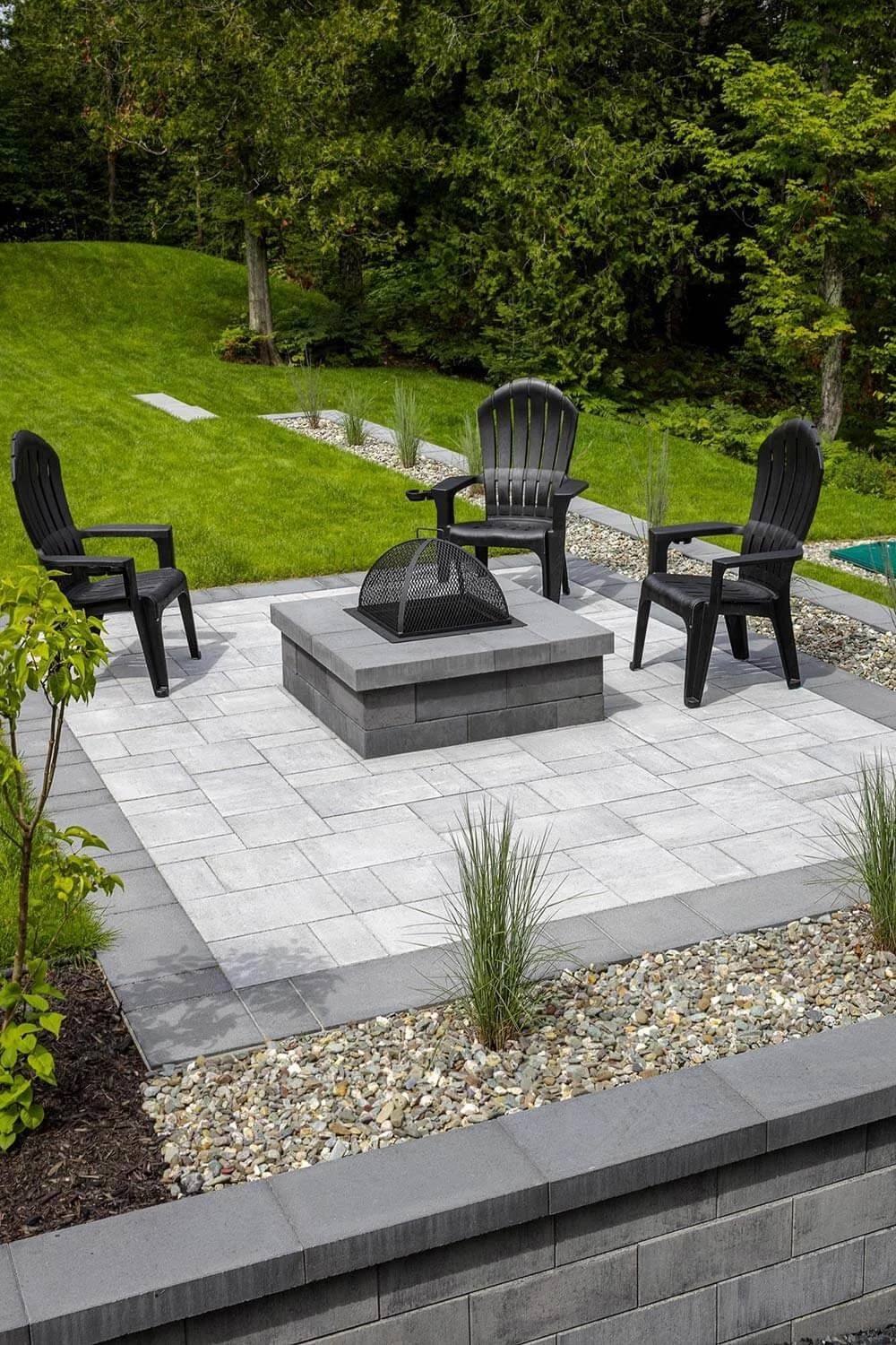 Outdoor patio with four black plastic chairs around a stone fire pit, surrounded by a neatly landscaped garden with grass, pebbles, and plants, bordered by trees in the background.