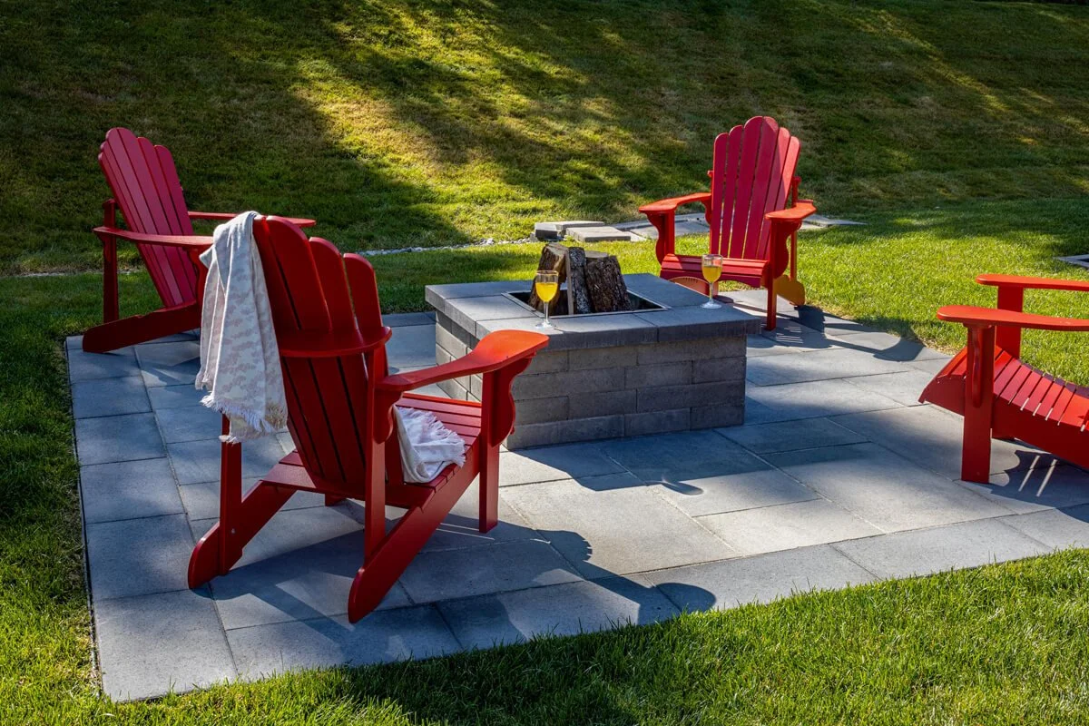 Outdoor patio with red Adirondack chairs around a stone fire pit, set on a paved area with green grass.