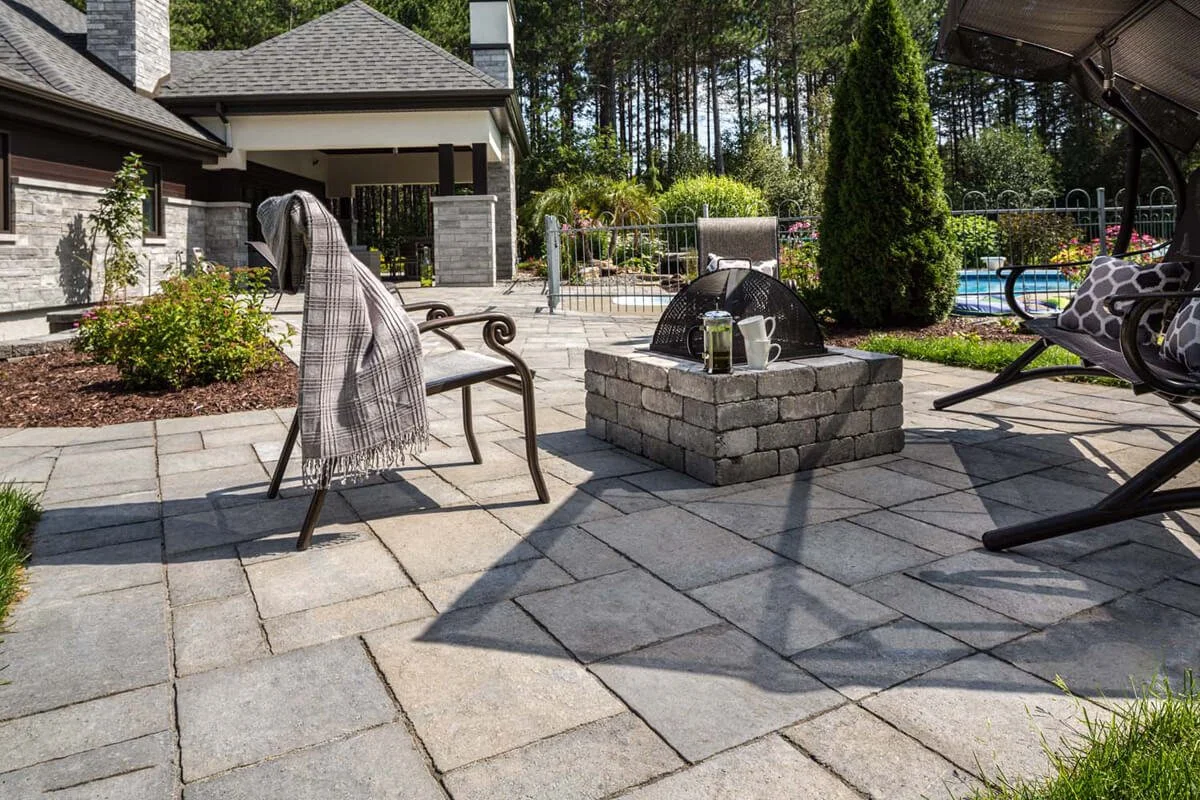 Outdoor patio with paving stones, seating area, fire pit, teapot on table, plaid blanket, garden and pool in background, surrounded by trees.
