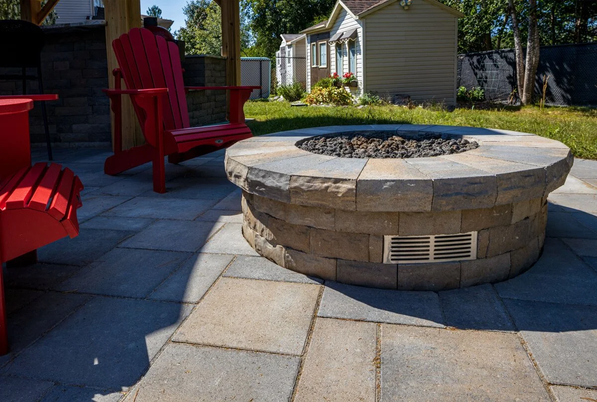 Outdoor patio with stone fire pit and red Adirondack chairs, set on a paved surface, with a small shed and greenery in the background.
