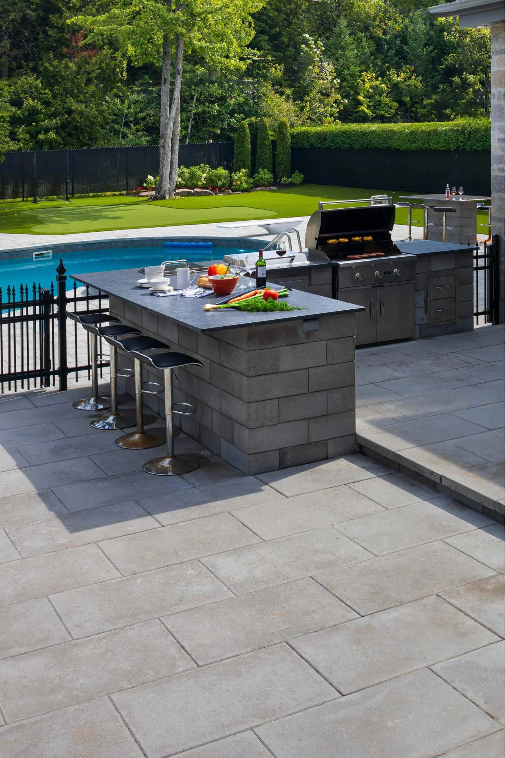 Outdoor patio with barbecue grill, stone countertop, bar stools, and pool in backyard setting.