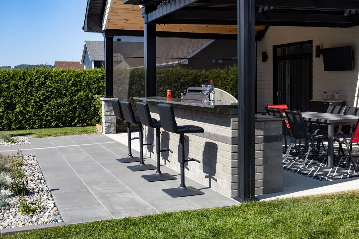 Outdoor patio with barstools and counter, adjacent dining area under a covered pergola, surrounded by green lawn and hedge.