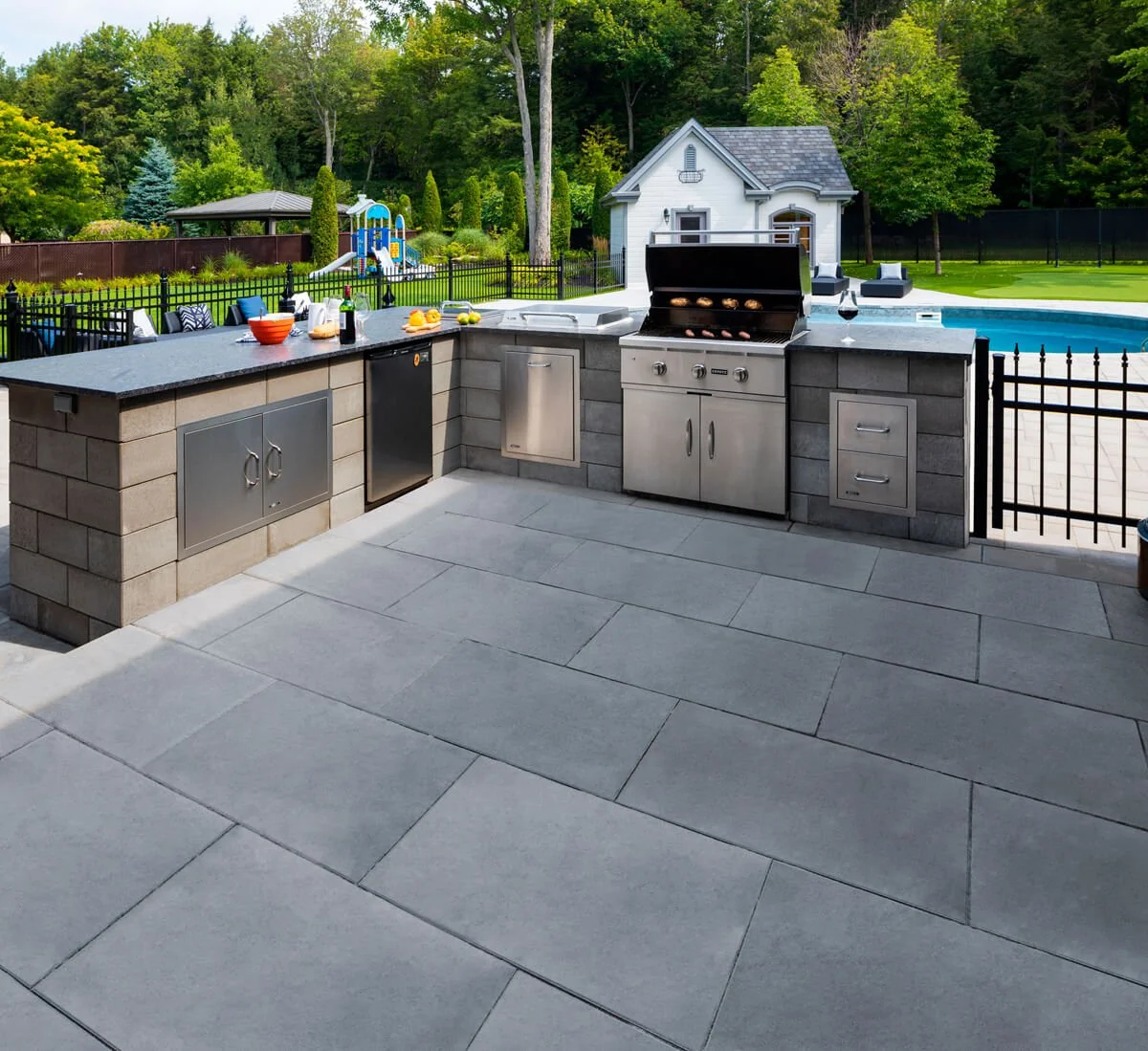 Outdoor kitchen with stainless steel appliances, including a grill and dishwasher, on a patio next to a swimming pool, surrounded by a black fence and overlooking a backyard with trees.
