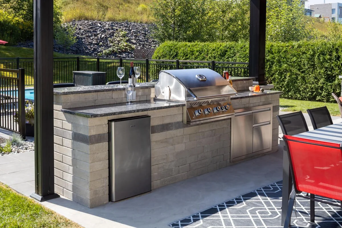 Outdoor kitchen with a built-in grill, granite countertop, mini fridge, and patio dining area.