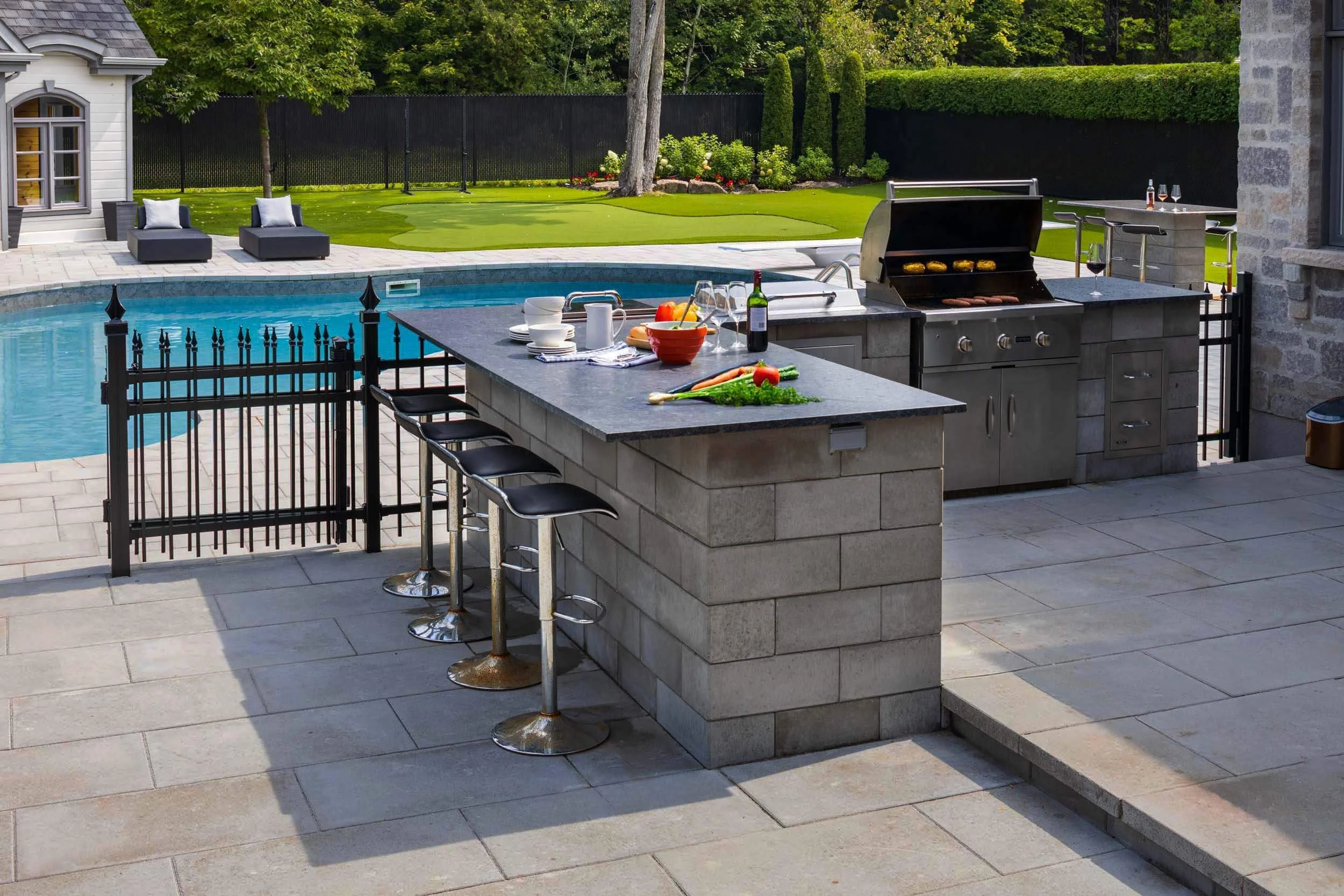 Outdoor kitchen area with a grill and countertop near a swimming pool, featuring bar stools and a small putting green in the background.
