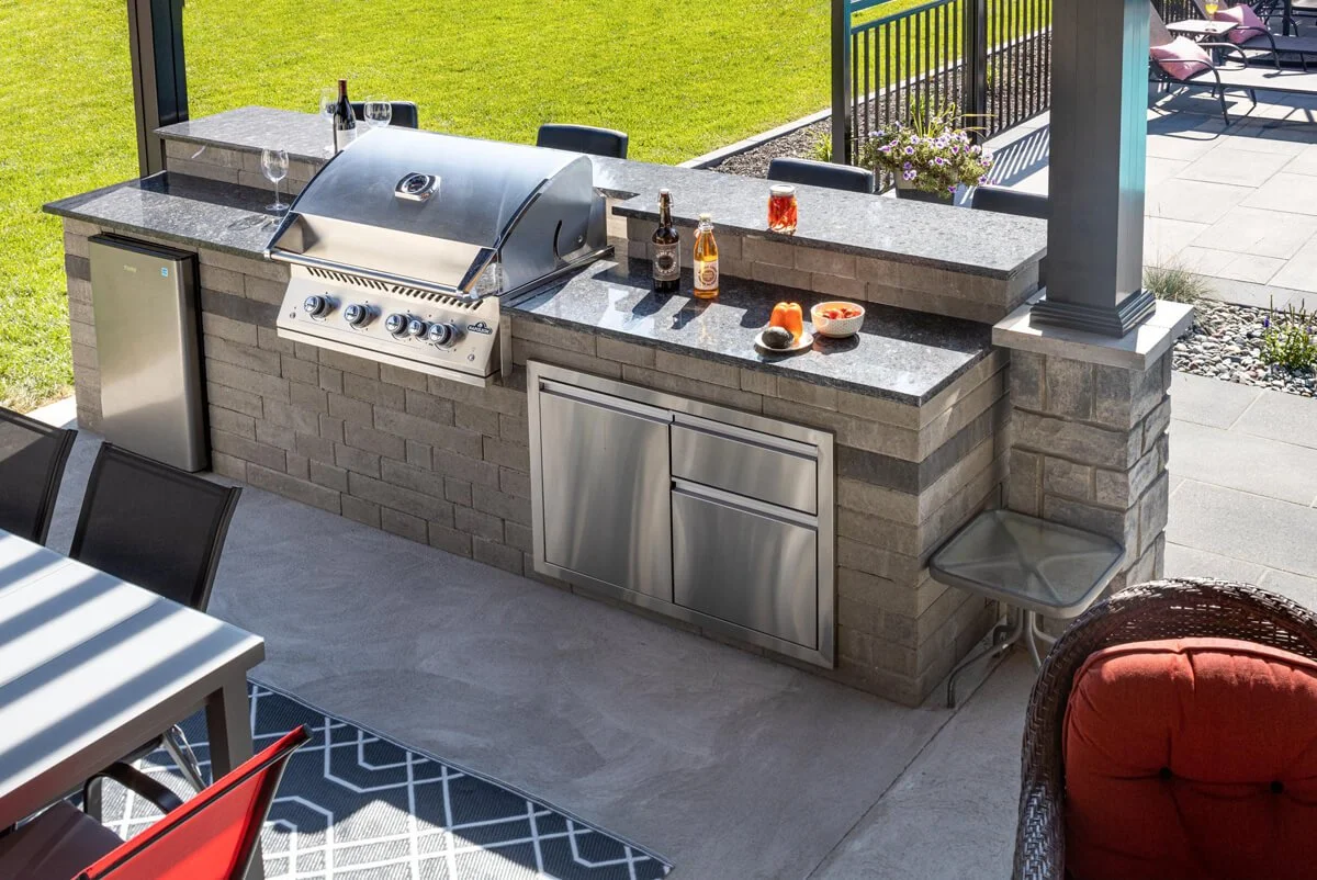 Outdoor kitchen with brick island, grill, mini fridge, and countertop seating, set in a backyard patio with grass and flowers.