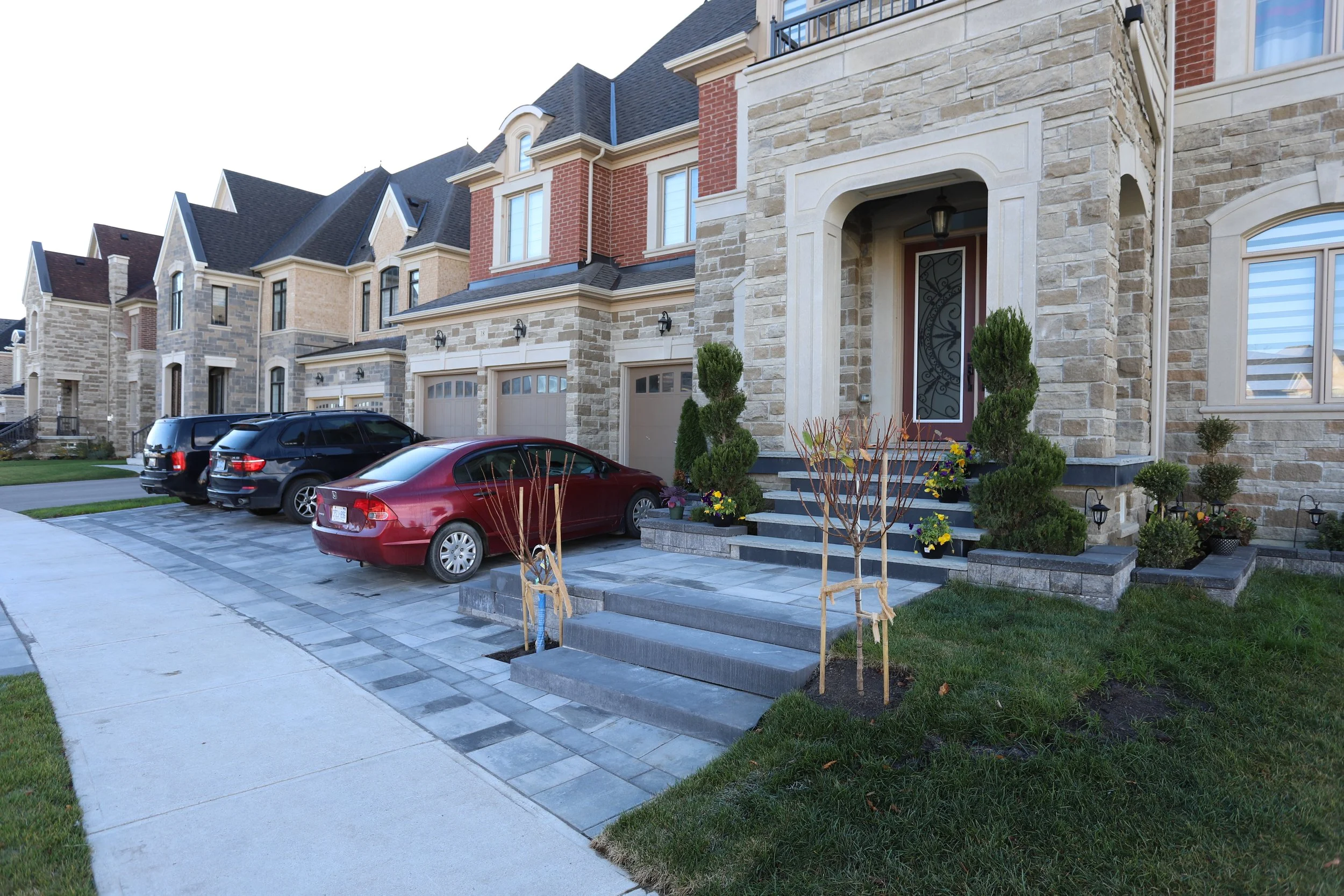 Suburban houses with stone facades and driveways, cars parked in front, steps leading up to an entrance, and landscaped gardens with small trees and shrubs.