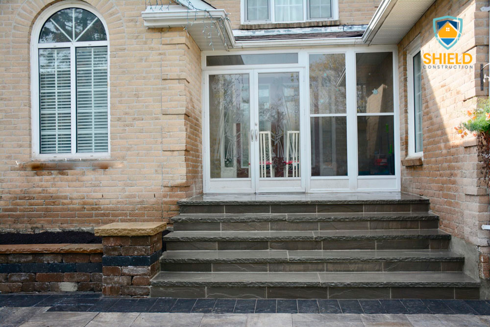 Exterior of a brick house with steps leading to a glass door entrance, featuring an arched window with white shutters on the left and 'Shield Construction' logo on the top right corner.