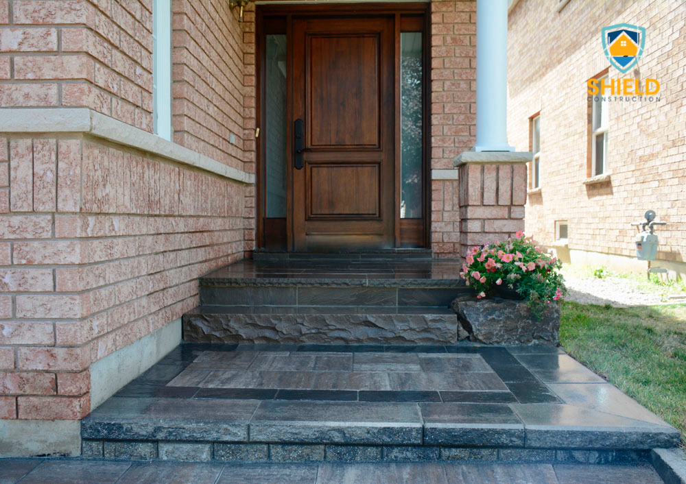 Brick house entrance with wooden door, tiled steps, potted pink flowers, and Shield Construction logo.