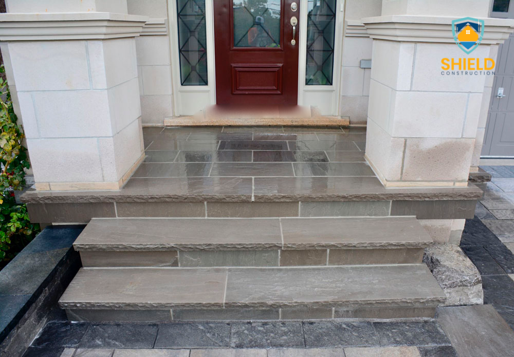 Stone steps leading to a front door with gray tiles and white columns. Entrance of a house with a dark red door. Shield Construction logo in the corner.