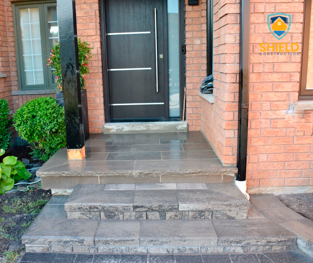 Front entrance of a house with stone steps and a modern black front door. The entrance features brick walls, a potted plant, and a glass window. A logo for Shield Construction is visible on the right side.