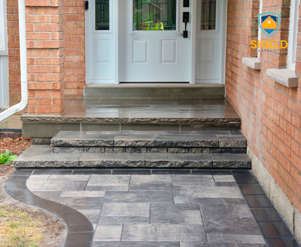 Front entrance of a house with brick walls and stone steps leading to a white door, featuring a paved walkway and a logo for Shield Construction in the corner.