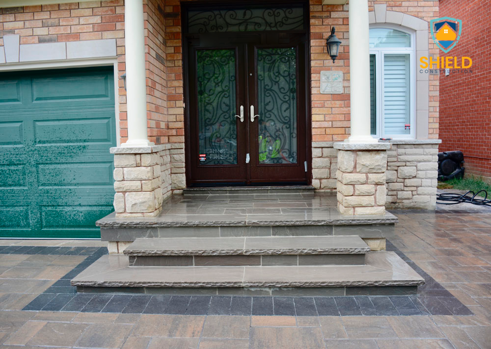Front entrance of a house with brick exterior, stone steps, and columns, next to a green garage door.