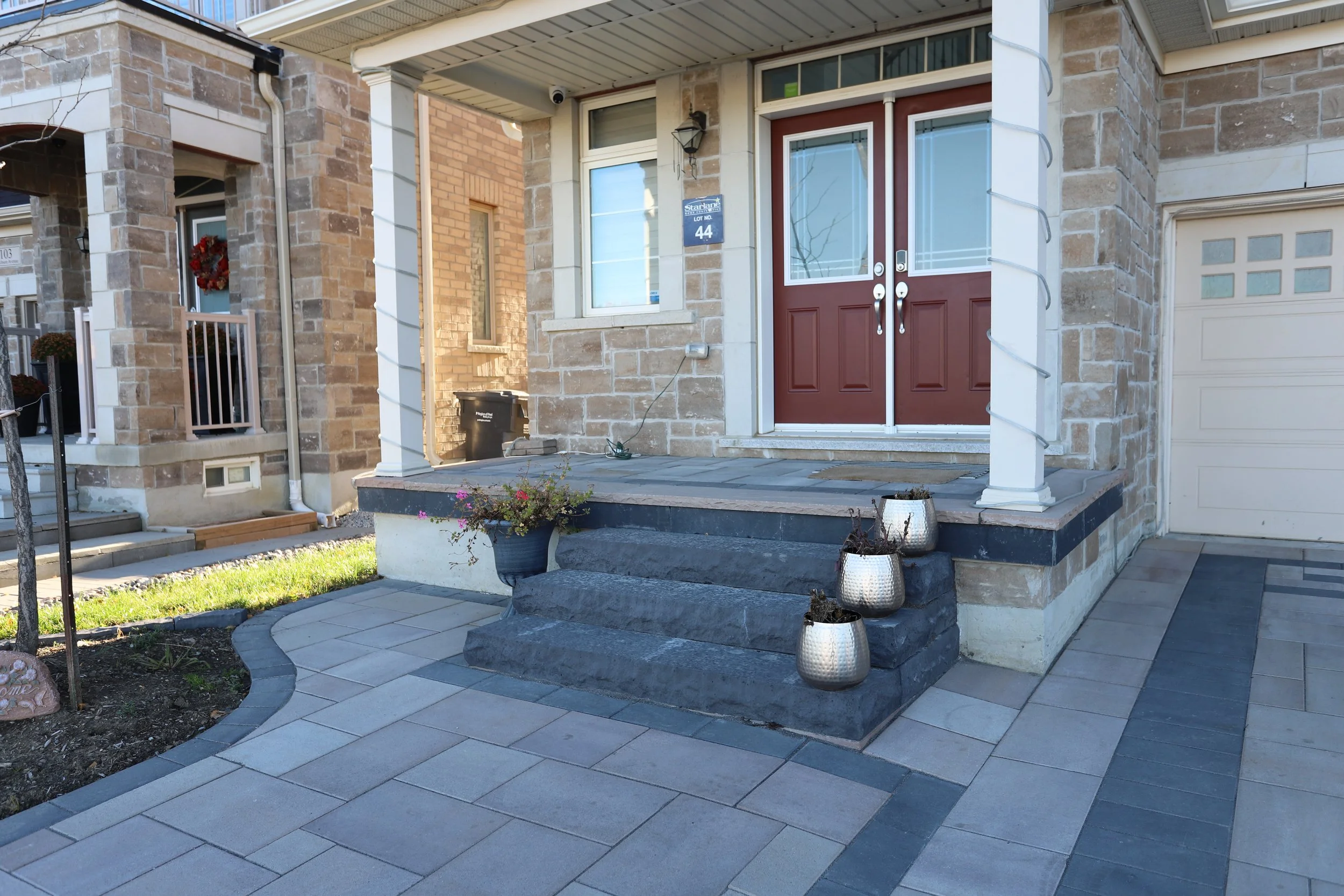 Front entrance of a house with double red doors, stone steps, and decorative planters on the steps. The house features stone facade and a paved walkway leading to the entrance.