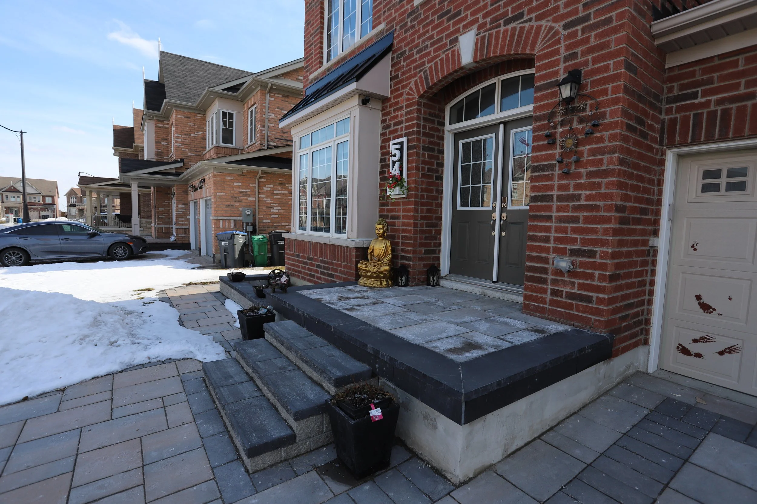 Front porch of a brick house with a gray door, a golden Buddha statue, and snow on the ground. There are black planters on the steps and a sidewalk leading to the driveway.