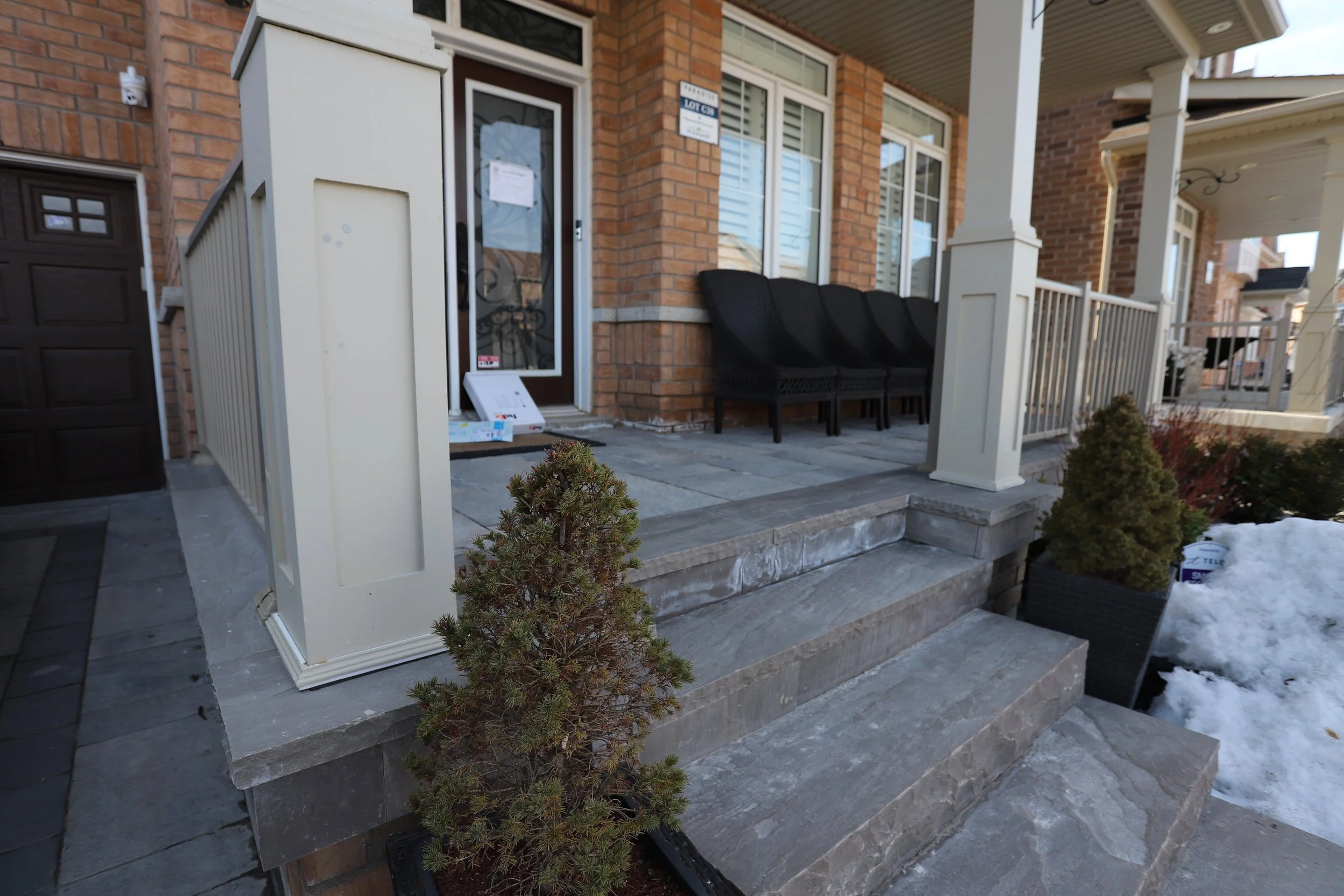 Front porch of a brick house with a few steps leading up to it. The porch has railing and wicker chairs. There are small evergreen shrubs in pots beside the steps, with snow visible on the ground nearby. A package is left at the front door.