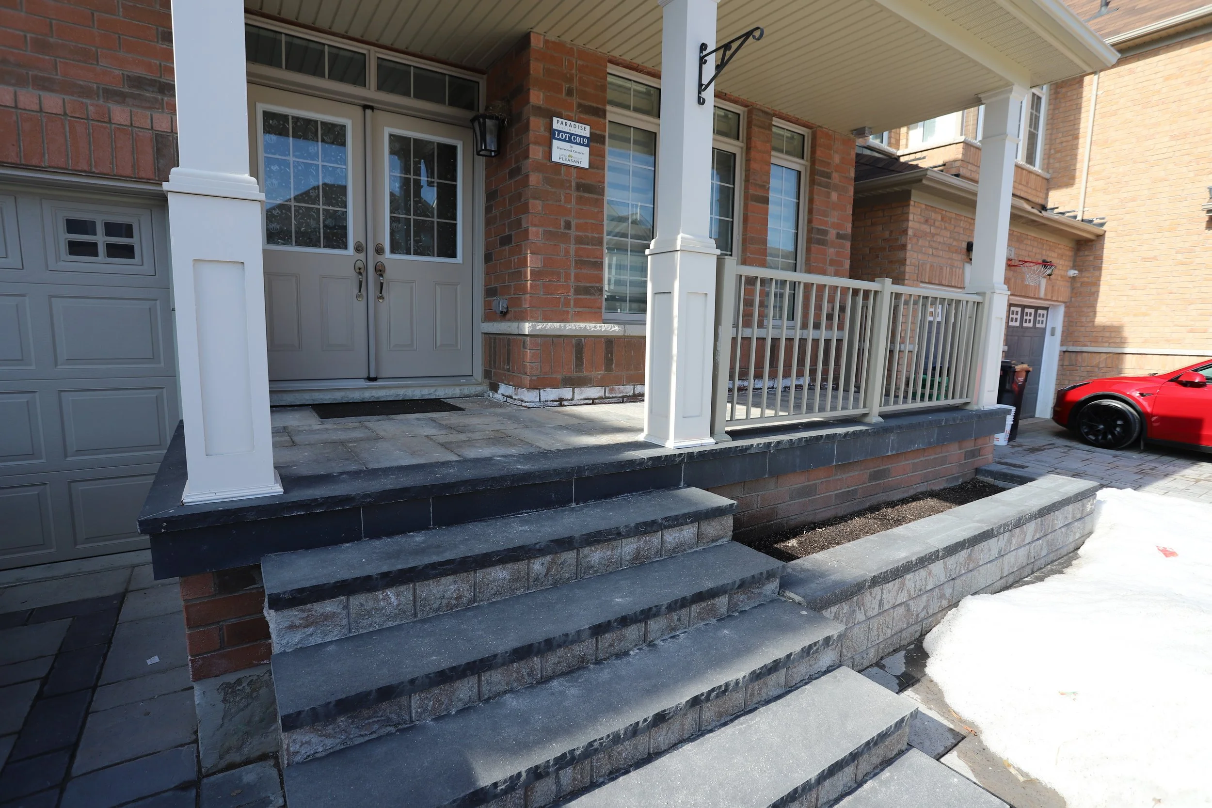Brick house entrance with double doors, stone steps, and adjacent driveway with a parked red sedan.