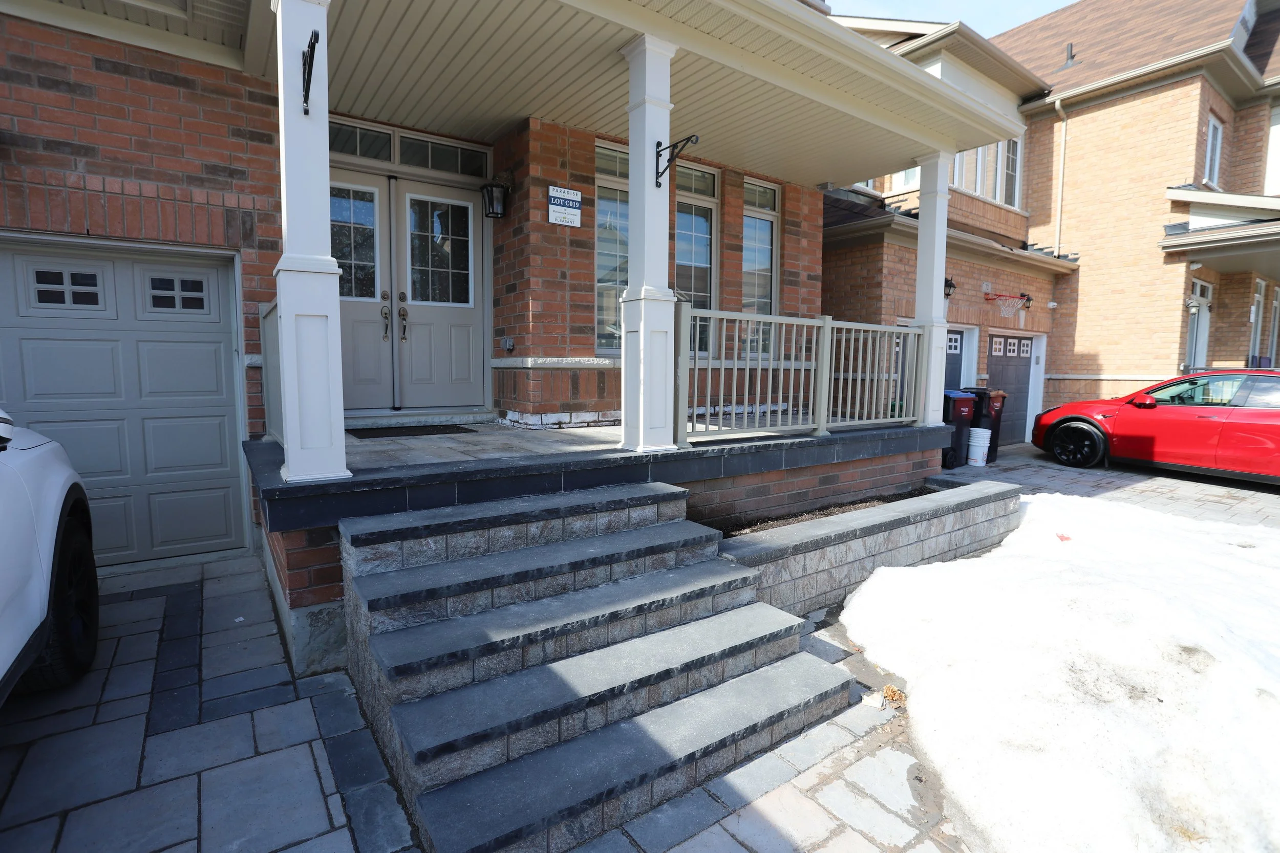 Front entrance of a brick house with steps leading to a porch, two garage doors, and a parked white car. A red car is parked in the driveway with snow on the ground.