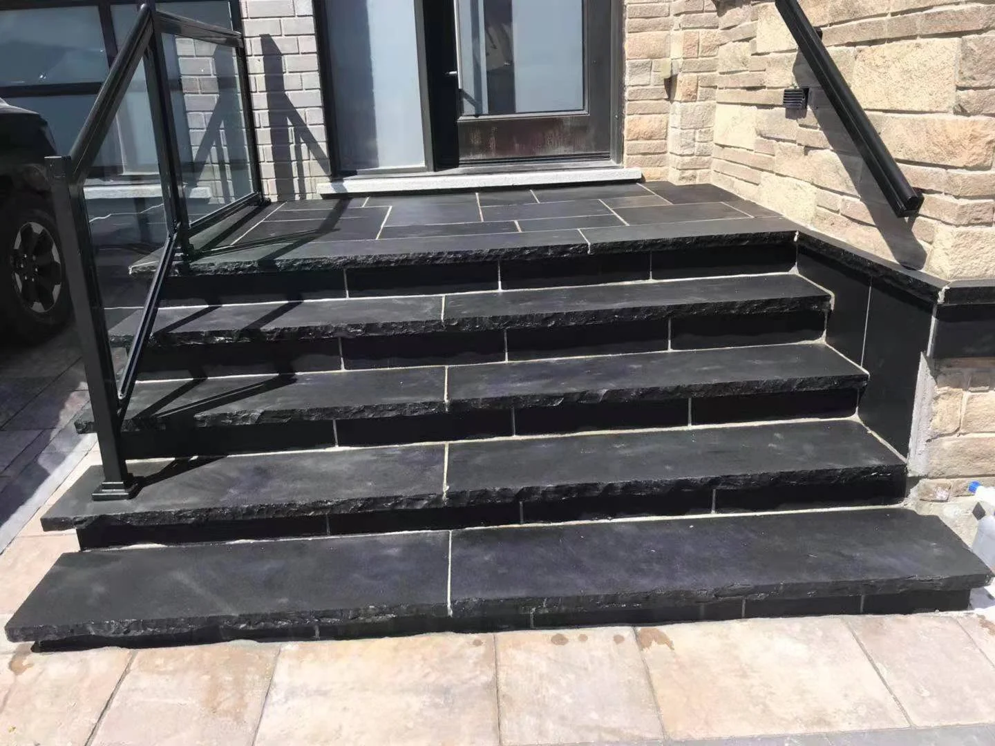 Black stone exterior stairs with glass railing leading to front door, surrounded by brickwork.
