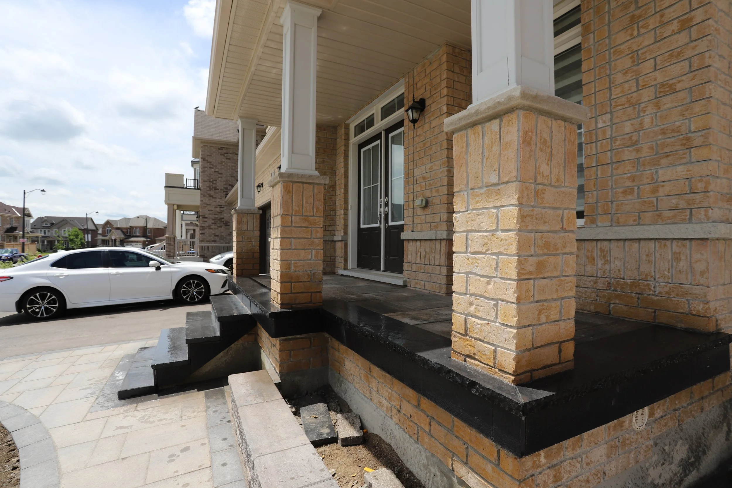 Front porch of a brick house with steps, columns, and a white car parked nearby in a residential neighborhood.