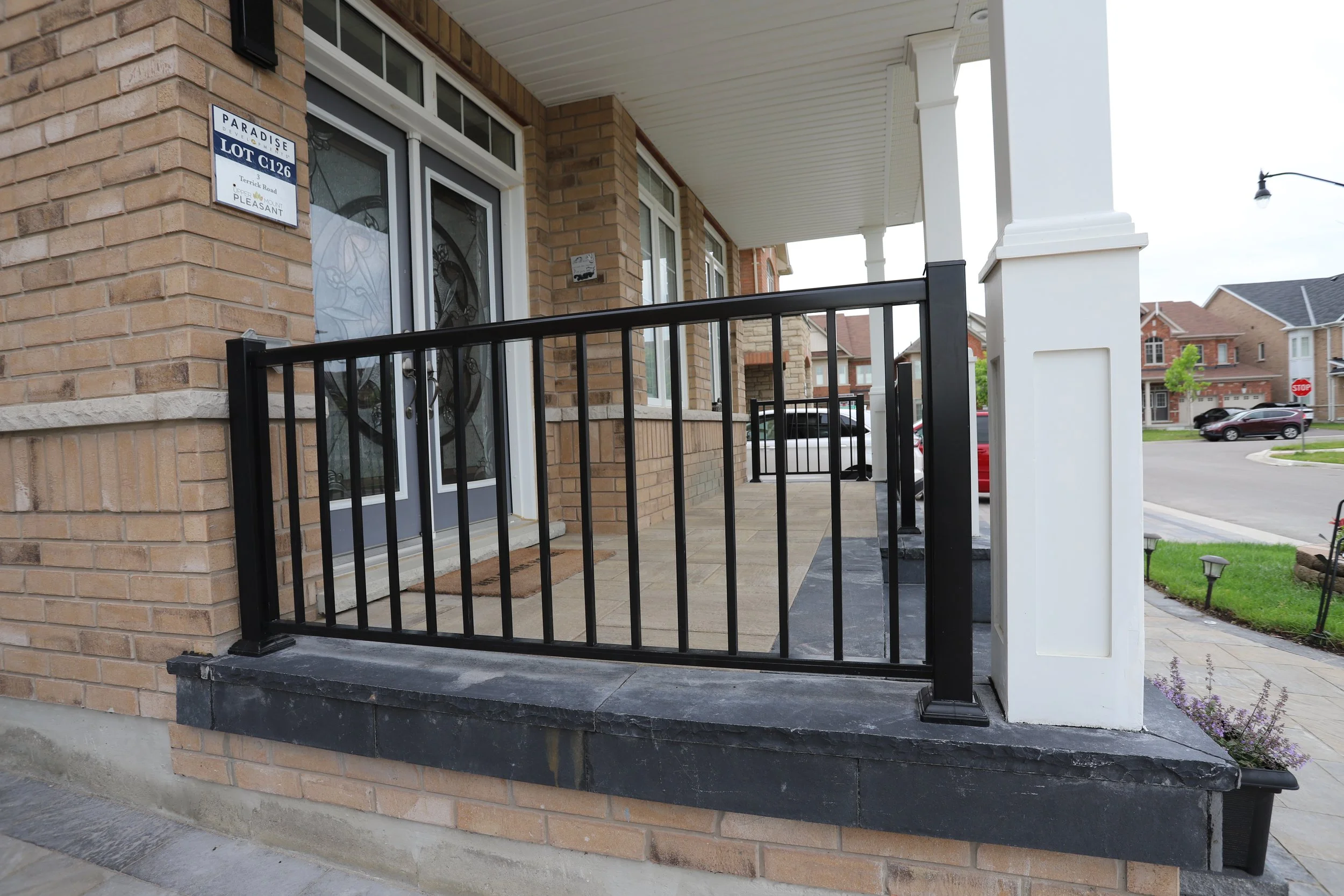 Front porch of a modern brick house with black metal railing and glass-paneled double doors, located in a suburban neighborhood.