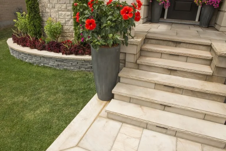 Front porch with stone steps and a planter with red flowers, surrounded by landscaped greenery and flower beds.