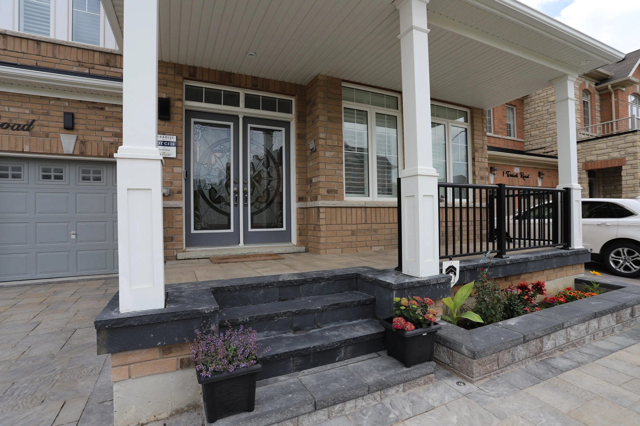 Front porch of a brick house with dual glass doors, a two-step entrance with black planters, and a nearby garage door. A white SUV is parked in the driveway.