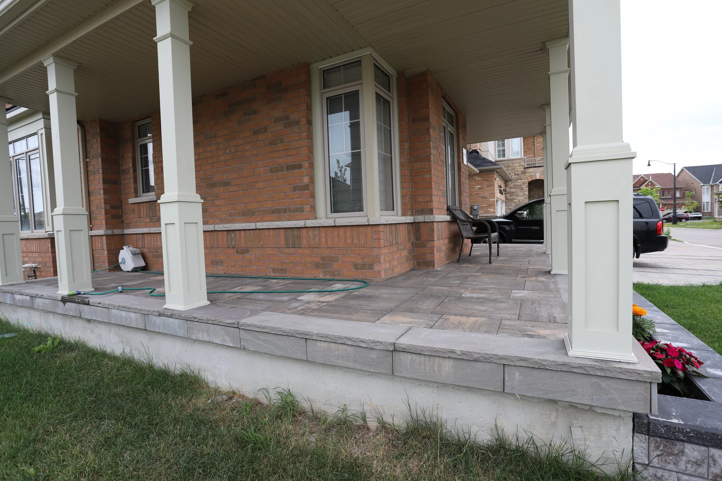 Residential porch with brick facade and columns, featuring a paved floor, outdoor seating, and garden hose, with lawn and driveway visible.