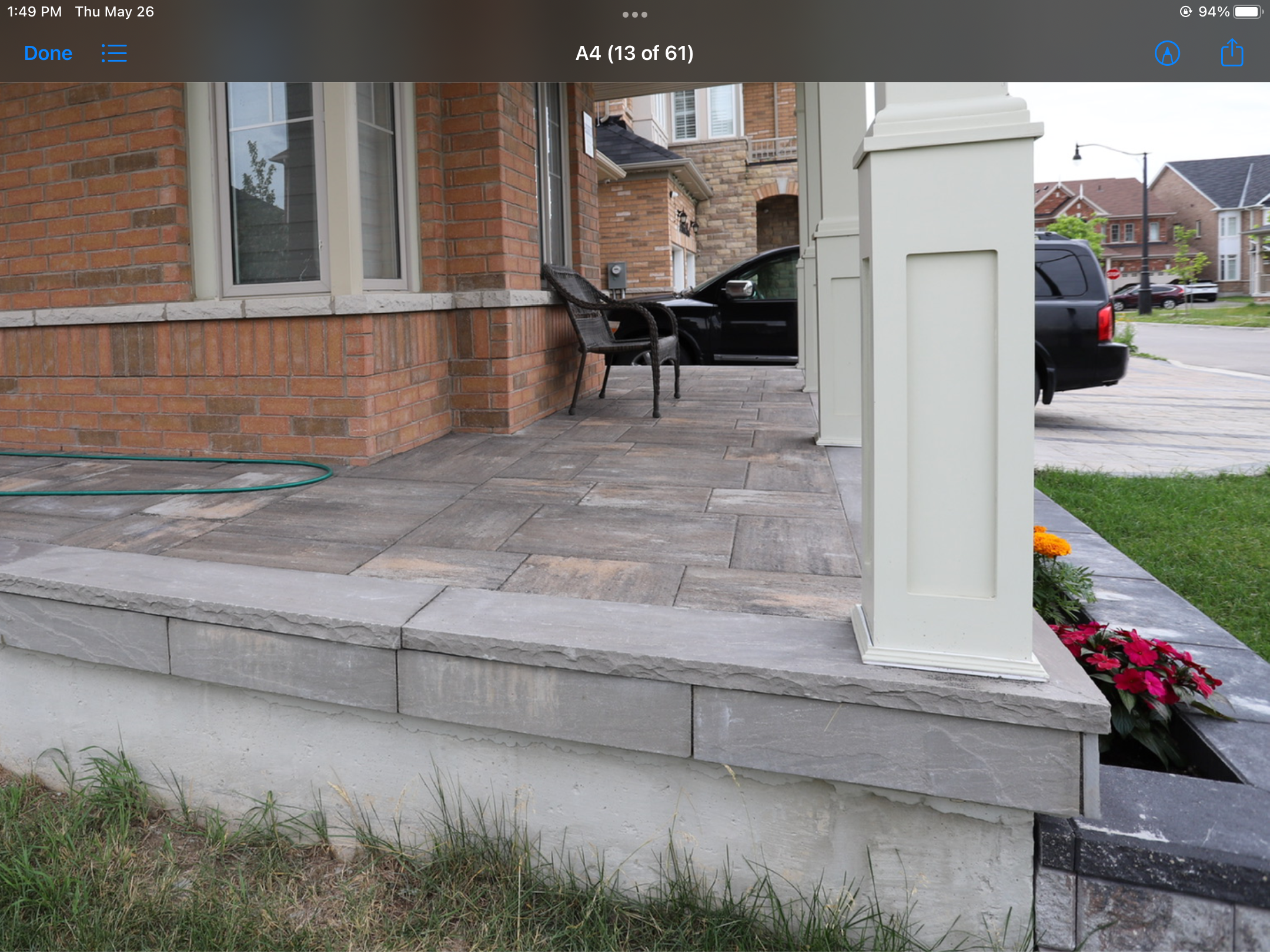 Front porch of a brick house with a tiled floor, a black chair, and a garden hose. A white column supports the porch roof. A black vehicle is seen parked on the driveway and a flower bed with red and orange flowers is visible.