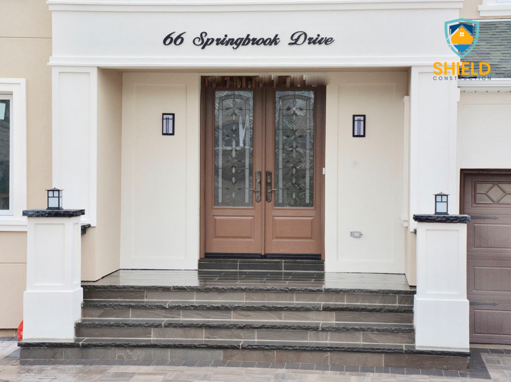 Front entrance of a modern house with double doors and address number 66 Springbrook Drive, featuring stone steps, decorative lanterns, and Shield Construction logo.