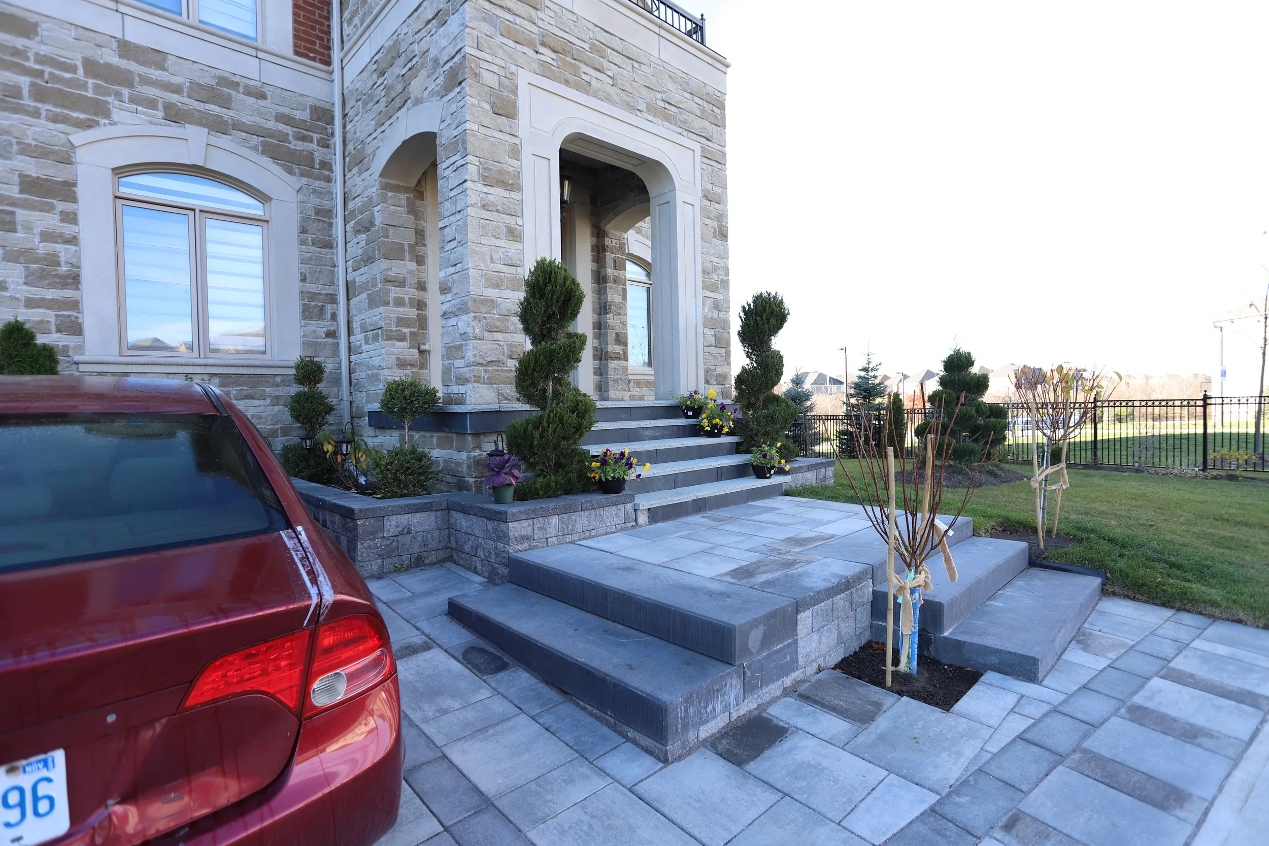 Front entrance of a house with stone facade, gray steps, potted plants, and a red car in the driveway.