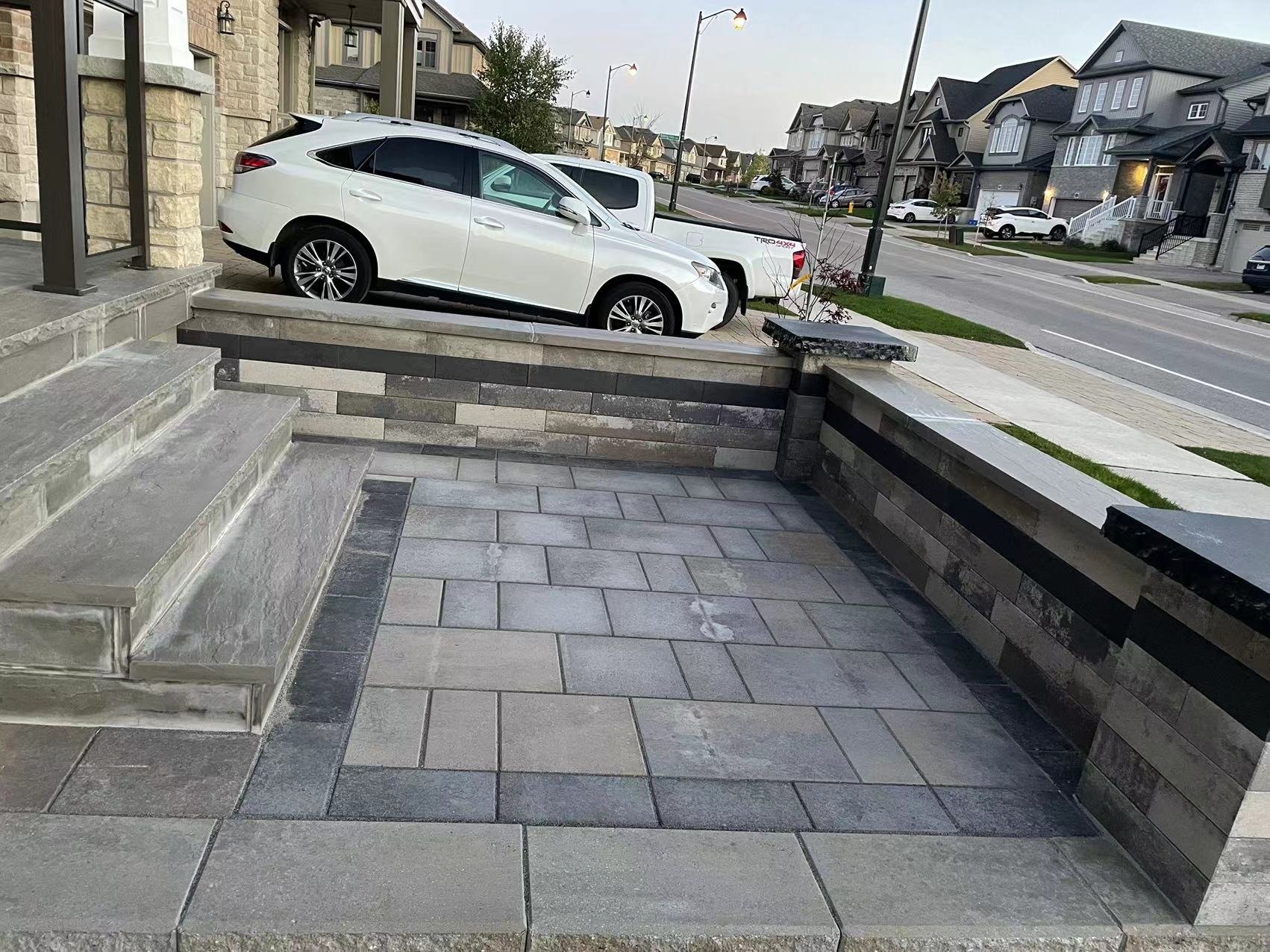 Residential street view with a white car parked in a driveway next to a house with stone steps and a paved patio. The neighborhood features multiple two-story homes.
