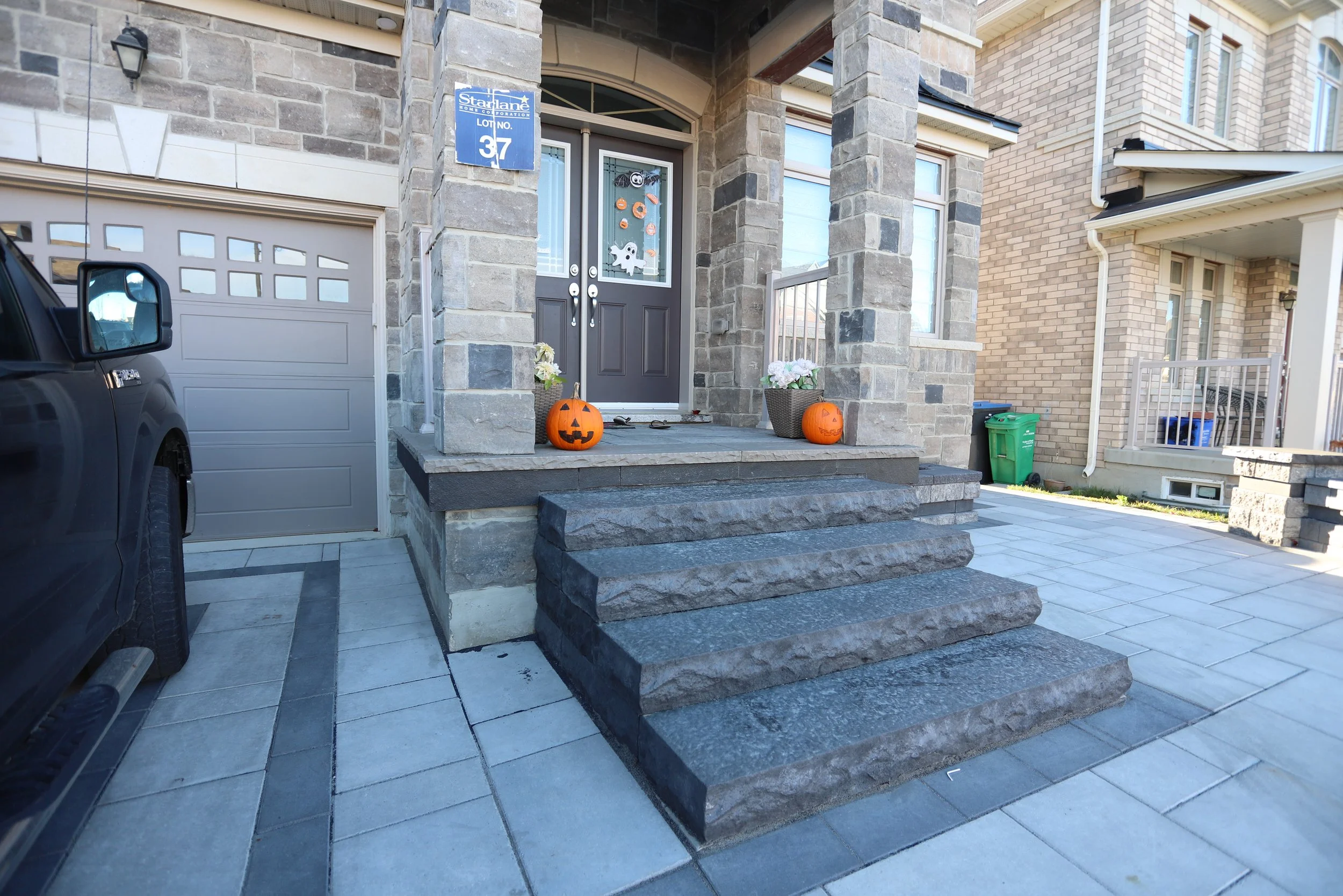 Front entrance of a house with stone porch and Halloween decorations, including two carved pumpkins with faces on the steps. A driveway with a black vehicle is visible, along with a garage door and recycling bins. The door has Halloween decals.
